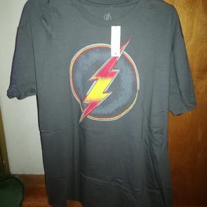 May include: A dark grey t-shirt featuring a yellow and red lightning bolt graphic within a circle. The graphic is a stylised version of the Flash logo.