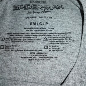 May include: A close-up of a grey t-shirt label featuring the "Spider-Man: No Way Home" logo. The label includes the Marvel Studios logo, size details, and fabric composition. Washing instructions and manufacturer information are also present.