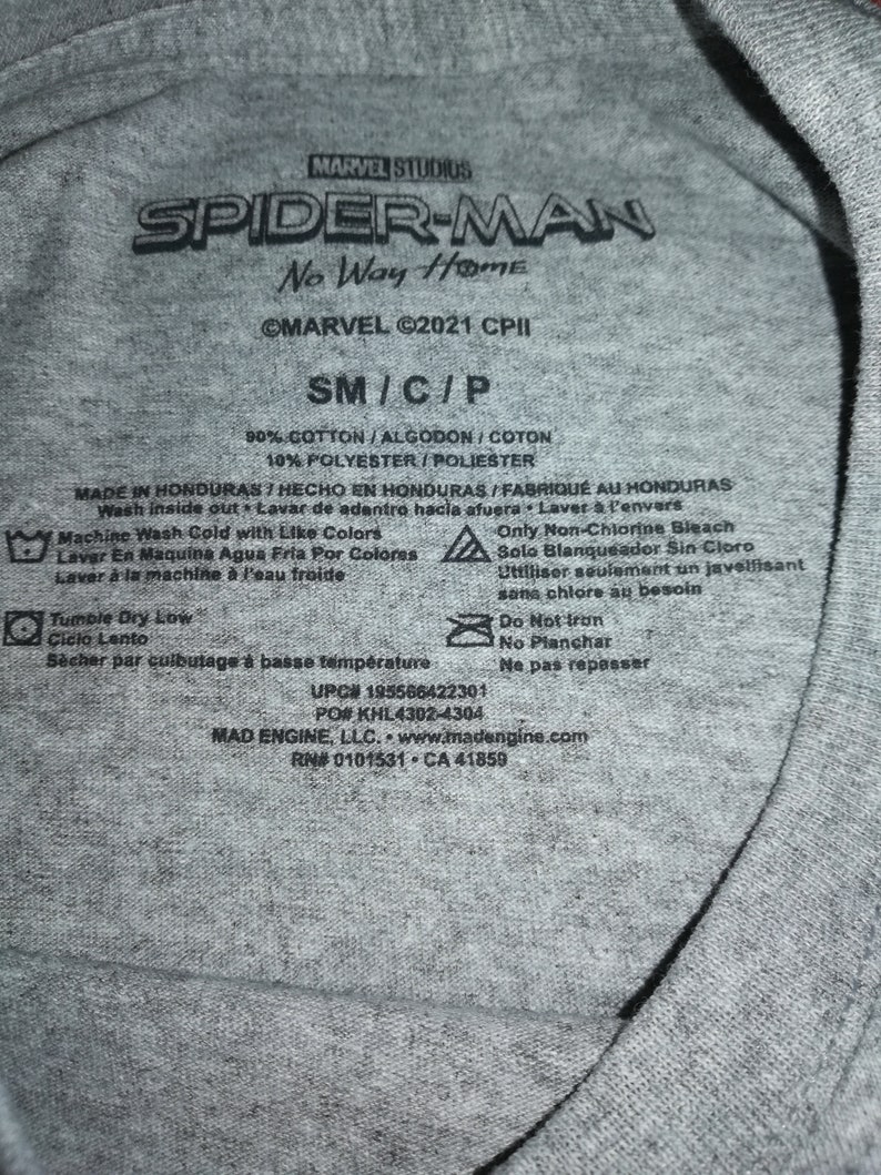 May include: A close-up of a grey t-shirt label featuring the "Spider-Man: No Way Home" logo. The label includes the Marvel Studios logo, size details, and fabric composition. Washing instructions and manufacturer information are also present.
