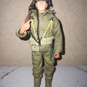 May include: A detailed action figure of a soldier in a World War II uniform. The figure wears a green helmet, olive drab jacket and pants, and brown boots. The figure is carrying a rifle and has a knife attached to the belt. The figure has a realistic face.