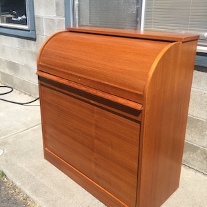 May include: A wooden roll-top desk with a warm, reddish-brown finish. The desk has a curved roll-top design and a rectangular base. The desk is made of wood and has a smooth, polished surface. The desk is a vintage piece of furniture.