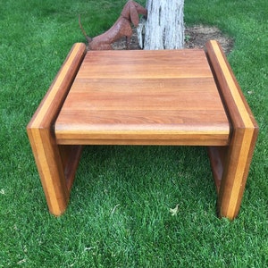 May include: A rectangular wooden coffee table with a natural wood finish. The table has a solid top and a frame with a contrasting wood stripe. The table is set on a green lawn.