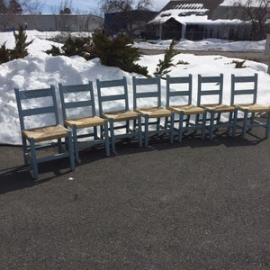 May include: A set of seven light blue wooden chairs arranged in a row. Each chair features a ladder-back design with a carved horizontal top rail and a woven natural fiber seat. The chairs have four straight legs connected by simple stretchers.