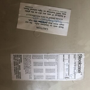 Puede incluir: Un primer plano de una etiqueta blanca con texto negro que dice "CAUTION" y "CLEAN CHAIR GLIDES PERIODICALLY". La etiqueta est&aacute; pegada a una silla beige con una etiqueta negra que dice "Steelcase" y tiene un n&uacute;mero de modelo y un n&uacute;mero de operador.