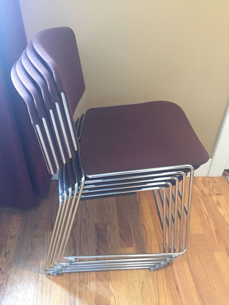 Puede incluir: Una pila de seis sillas de tela marr&oacute;n con marcos cromados. Las sillas est&aacute;n apiladas ordenadamente, con los asientos hacia afuera.