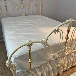 May include: Cream-colored metal bed frame with ornate detailing. The headboard and footboard feature decorative scrollwork and gold-colored accents. The bed is made up with a white fitted sheet and a white bed skirt.