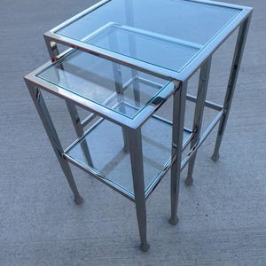 May include: A set of two nesting glass top tables with chrome frames. The tables are square and have a modern design.
