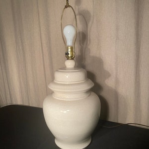 May include: A white ceramic table lamp with a textured finish. The lamp has a round base and a gold-colored metal shade.