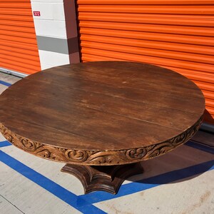 May include: A round, dark brown wooden table with a carved decorative edge. The table has a pedestal base and is approximately 48 inches in diameter. The wood grain is visible on the tabletop.