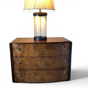 May include: A three-drawer chest featuring a curved front with a brown burl wood veneer, dark brown side panels, and small circular brass pulls. A table lamp with a cylindrical glass base and a beige fabric shade sits on the top surface.
