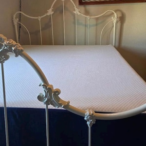 May include: A white metal bed frame with ornate floral detailing on the headboard and footboard. The bed has a white mattress and a navy blue bed skirt. The headboard has a curved design.