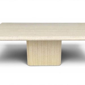May include: A square, beige travertine coffee table. The table has a solid, rectangular base and a smooth, flat top. The edges of the table are rounded. The table is approximately 48 inches long and 48 inches wide.