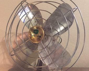 May include: A vintage Coast-Air electric fan featuring four metallic blades protected by a circular wire cage. The center of the fan has a gold-toned circular emblem with the text Coast-Air. The base is off-white with visible signs of wear.