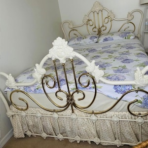 May include: A white metal bed frame with gold accents and ornate detailing. The bed is made up with a floral patterned comforter and pillows. The bed skirt is a cream color with a floral pattern.