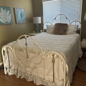 May include: White metal bed frame with a white and gold headboard and footboard. The bed is made with a white bedspread and a fluffy brown throw pillow.