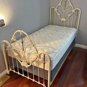 May include: White metal bed frame with decorative floral accents and brass-colored details. The bed has a mattress and box spring. The headboard and footboard feature ornate designs. The bed is on a hardwood floor.