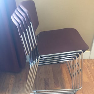 Puede incluir: Una pila de seis sillas con asientos de tela marr&oacute;n y marcos cromados. Las sillas est&aacute;n apiladas de forma ordenada, con los asientos mirando hacia afuera.