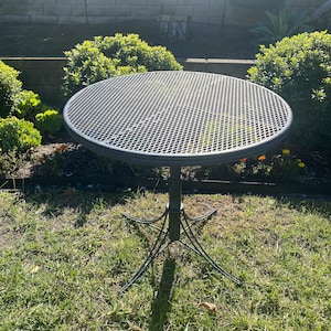 May include: Black metal patio table with a round, wire mesh top and a tripod base. The table is sitting on a grassy lawn.