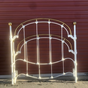 May include: White metal bed frame with gold accents. The frame has a decorative design with curved lines and floral details. The bed frame is in front of a red garage door.
