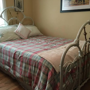 May include: A white metal bed frame with a plaid comforter in red, green, and white. The bed has two pillows and a decorative throw. The headboard has ornate detailing.