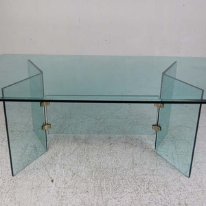 May include: A rectangular glass dining table with a clear glass top and a unique, geometric base made of four glass panels connected by brass hinges. The table has a modern, minimalist design.