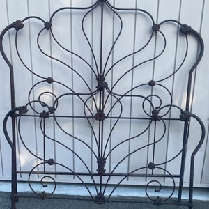 May include: Ornate, dark metal bed frame with a decorative headboard. The headboard features a symmetrical design with curved lines and floral accents. The frame is made of wrought iron and has a vintage aesthetic.