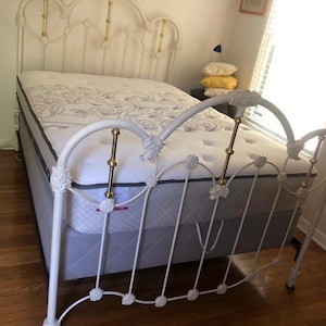 May include: A white metal bed frame with a decorative headboard and footboard. The bed has a mattress with a patterned design and a gray box spring. The frame has gold accents and is set on a hardwood floor.