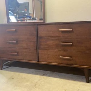 May include: A six-drawer dresser with a dark wood finish and simple, modern hardware. The dresser has a mid-century modern style.