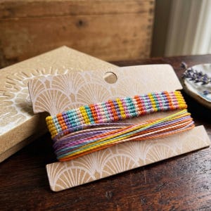 Könnte beinhalten: Zwei farbenfrohe Freundschaftsarmbänder auf einer dekorativen Karte, mit einer passenden Geschenkbox im Hintergrund. Die Armbänder weisen eine Vielzahl von Farben auf, darunter Orange, Gelb, Blau und Lila. Die Geschenkbox ist hellbraun.