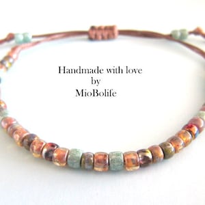 Può includere: Un braccialetto delicato realizzato con piccole perline colorate in tonalità di marrone, arancione, rosso e verde. Il braccialetto è legato con un cordoncino marrone e ha un nodo all'estremità. Il testo "Handmade with love by MioBolife" è visibile nell'immagine.