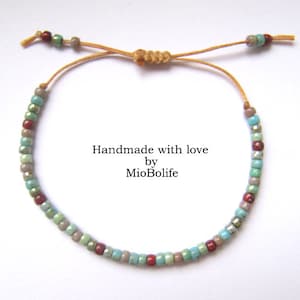 Può includere: Un braccialetto fatto a mano con perline turchesi, grigie e rosse su un cordoncino marrone. Il braccialetto è legato con un nodo e ha un'etichetta che dice "Handmade with love by MioBolife".
