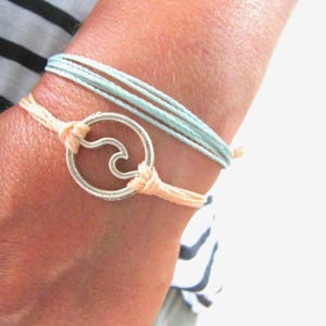 Può includere: Un primo piano di un polso che indossa due braccialetti. Il braccialetto superiore è blu chiaro e il braccialetto inferiore è pesca con un ciondolo a forma di onda argentato.