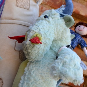 May include: A blue and white plush toy dragon with a red tongue and a small doll wearing a blue and white shirt riding on its back.