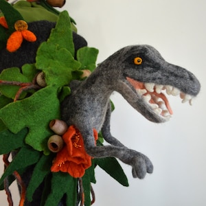 May include: A gray felt dinosaur with an open mouth and orange eyes peeking out from behind green felt leaves.