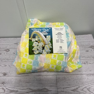 May include: A fitted crib sheet with a checkered pattern in yellow, blue, and white, featuring small teddy bear, block, and rattle icons. A product label titled Teddy Time Collection is attached, showing a rainbow and listing 100% cotton material.
