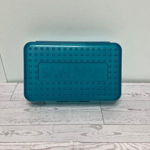May include: A rectangular, translucent teal plastic pencil case with a hinged lid. The surface features a grid of small, raised circular bumps and the embossed text "Spacemaker" centered on the top cover of the storage container.