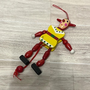 May include: A wooden marionette doll with a painted face, red limbs, and a yellow torso. The doll's arms and legs are jointed, and it has black feet. A red string is attached to the doll for manipulation.