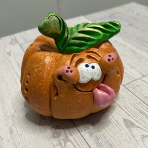 May include: A round orange pumpkin figurine with a textured surface, featuring a large green leaf on top. The pumpkin has a cartoonish face with two white eyes, pink cheeks, and a pink tongue sticking out from a wide, open mouth expression.