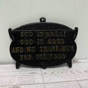 May include: Black cast iron decorative pot-shaped sign with the words "GOD IS GREAT GOD IS GOOD AND WE THANK HIM FOR OUR FOOD" in gold lettering. The sign has a loop at the top for hanging and three small feet.