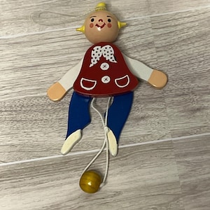 May include: A wooden pull-string toy doll with a painted face, yellow hair, and a red vest with white accents. The doll has blue pants, white arms, and a yellow ball at the end of the string. A classic children's toy.