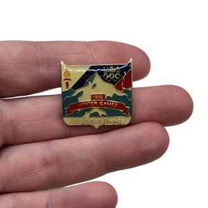 May include: A gold-colored pin with a white, blue, and red design. The pin features a mountain range, a torch, and the Olympic rings. The text "1992 Winter Games Albertville France" is printed on the pin.