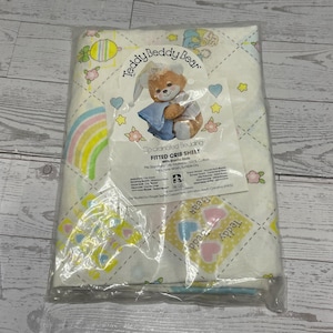 May include: A white fitted crib sheet featuring a Teddy Beddy Bear pattern with rainbows, hearts, and flowers. The packaging includes the text Teddy Beddy Bear, Co-ordinated Bedding, FITTED CRIB SHEET, 100% Cotton, and details from the Riegel Textile Corporation.