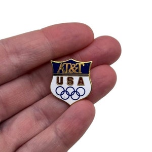 May include: A gold-colored pin with a white shield-shaped design. The shield has a blue band at the top with the text "AT&T" in gold letters. Below the band, the text "USA" is in gold letters. The bottom of the shield features the Olympic rings in blue.