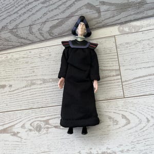 May include: A small figurine of a character in a black robe with a tall, pointed hat. The figure has a pale face and is standing on a light-colored wooden surface. The hat is black and blue.