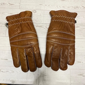 May include: A pair of brown leather gloves with white stitching. The gloves have a textured appearance and a zipper detail on the side. The gloves are designed for warmth and protection.