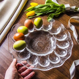 May include: A clear glass egg plate with 12 individual compartments for holding eggs. The plate has a scalloped edge and a raised centre. There are four green and brown eggs in the plate.