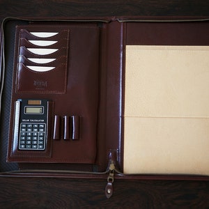 May include: A brown leather zippered folder with a calculator, pen holder, and card slots. The folder has a cream-colored interior.