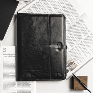 May include: Black leather portfolio with a strap and the word "NOIR" embossed on the front. The portfolio is on a white surface with a magazine and a pen.