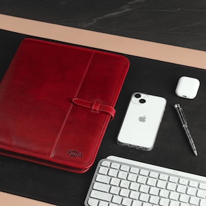 May include: A red leather portfolio with a black strap and the word "Nood" embossed on the front. The portfolio is on a black desk mat with a white wireless keyboard, a white iPhone, a white AirPod case, and a black pen.
