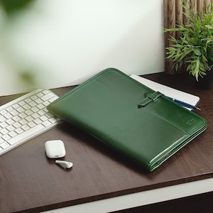 Puede incluir: Un portafolio de cuero verde oscuro con un teclado blanco, auriculares blancos y un cuaderno con un bolígrafo sobre un escritorio de madera oscura.